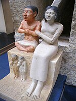 statue partiellement peinte d'un petit homme vêtu seulement d'un pagne, assis en tailleur à côté d'une femme vêtue d'une robe longue