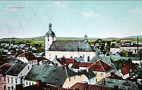 Kolorovaná pohlednice, uprostřed kostel s věží vlevo a presbytářem vpravo, v popředí a po stranách městská zástavba, v pozadí obdělávané kopce a obloha