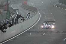 Photographie d'une voiture bleue, blanche et rouge, de face, sur une piste, devant un drapeau à damier