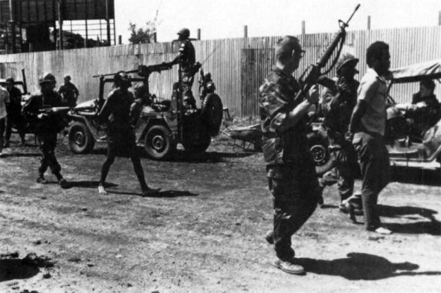 Two men in civilian clothes with their hands on their backs walk surrounded by three armed men in uniform. Military jeeps are seen in a second plane.