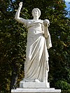Statue von Urania im Park Sanssouci, Potsdam, Frontalansicht; ihr rechter Arm ist über ihr erhoben, die Finger zusammengebogen, eventuell war dort einmal ein Stab hineingesteckt; ihr linker Arm ist halb nach vorn ausgestreckt, in der Hand hält sie eine Kugel