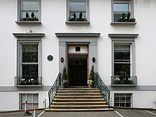 A flight of stone steps leads from an asphalt car park up to the main entrance of the white two-story building. The ground floor has two sash windows, the first floor has three shorter sash windows. Two more windows are visible at basement level. The decorative stonework around the doors and windows is painted grey.