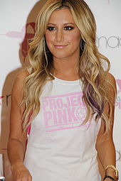 A young female with long blonde hair wearing a white t-shirt with "PROJECT PINK" written on it smiles to the camera.