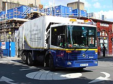 A large six-wheeled lorry operated by the "City of Westminster", with a waste disposal unit at rear, drives across a small roundabout at the corner of a busy junction