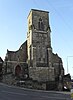 A dull-coloured pale stone church on a sloping corner site, with a tower stopping abruptly at a parapet.