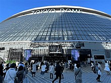 Menschen vor dem dem Eingang zum Tokyo Dome, unter dem gläsernen Vordach ein Großplakat, das die Show "GIFT" ankündigt