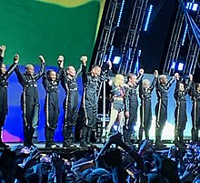 Image of several people raising their fists in front of a stage. The majority of them are wearing black jumpsuits, while a blond woman is wearing a different black outfit, with colorful stripes on her chest. She is also holding a microphone to her mouth.