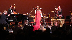 In the forefront is a crowd of people facing a stage. In the background are four people on a stage; a woman in the center is wearing a red dress and standing behind a microphone and the rest are men performing various instruments, including a piano, double bass and drum set.