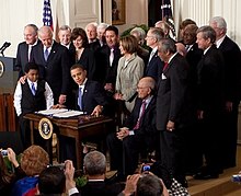 A man in a suit and tie sits at a wooden desk with two pieces of paper spread out in front of him. Behind him is a group of similarly dressed men and women; he is flanked by two young boys