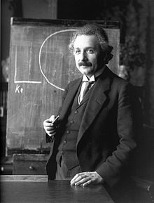 Einstein mit Kreide in der Hand vor einer Tafel stehend, auf die er einen Kreidekreis gezeichnet hat, lächelnd, von halblinks, die linke Hand auf dem Pult abstützend