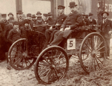 Photo d'une automobile avec un chauffeur et un copilote que regarde la foule.