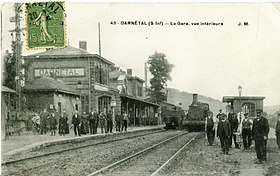 Ancienne carte postale montrant le bâtiment voyageurs et les quais (sur lesquels se trouvent des voyageurs et le personnel de la gare), avec deux trains.