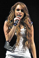 A brunette teenager sings into a microphone. She is wearing a black leather vest over a white tank top and shorts.