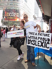 A girl holding a sign that says "LD = equally intelligent / Cross out stigma" poses for a photo in Times Square with a man holding a sign that says "Take a picture with a proud Dyslexic".