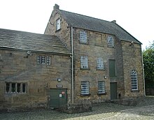 Worsbrough Mill on a bright day. The new mill is visible on the right, and the old mill abuts the new mill on the left. Both buildings are made from the same brown stone, and have the same stone roof; the new mill is higher. The new mill has three floors, and a total of 8 windows, one of which is a bottle window facing forward on the right, and one of which overlooks the old mill, facing left. The remaining six windows, facing forward, are arranged as follows. Ground floor: two adjacent windows. First floor: two adjacent windows, directly above the lower ones. Second floor: one window above the leftmost of the pair below, and one to the right of the rightmost window below. Directly above the rightmost window on the second floor is a false window that is apparently bricked in. The Old Mill has two windows visible and these are small and barred. Doors: there are three visible doors, one double door to the old mill, and two (one above the other) to the new mill, to the left of the bottle window. All doors are painted a dull green. There are two sloping stone structures in the foreground, about three feet high, to facilitate loading flour on to a wagon