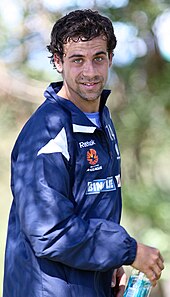 Alex Brosque, wearing a navy tracksuit top and a light blue shirt, holding a bottle of water
