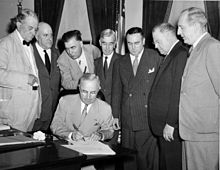 Un homme en costume est assis et signe un document. Sept autres hommes en costumes sont rassemblés autour de lui.