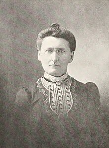 B&W portrait photo of a middle-aged woman with her hair in an up-do, wearing a dark blouse with a frilly front.