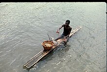 Photographie d'un homme, torse nu, assis à califourchon sur une planche en bois flottant sur l'eau. L'homme tient un filet contenant plusieurs poissons.