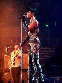 A female with medium skin and short black hair. The female is holding onto a microphone and a microphone stand with both her hands and is wearing black gloves that cover both her arms from above her elbow down. She is wearing black thigh high boots.