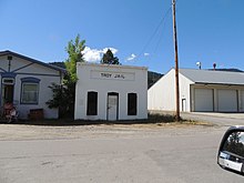 one story whitewashed concrete jail on a road. it has the words "troy jail" painted in block letters above the rectangular door