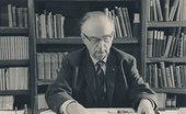 Older Hendrik Coenraad Blöte working at his desk. Hendrik is wearing glasses. a suit, tie and jacket. Many reference books can be seen on shelves in the background.