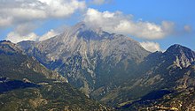 A rocky peak partly covered in cloud, behind lower tree-clad mountains