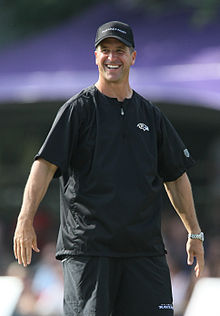 Knees-up photograph of Harbaugh smiling while wearing a dark Baltimore Ravens t-shirt and a Ravens baseball cap