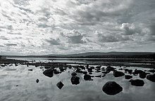 Pohled na jezero, vepředu desítky balvanů, vzadu les a kopce