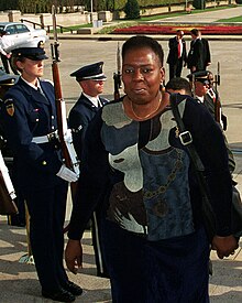 Une photographie en couleurs d'une femme noire montant un escalier qu'entourent des militaires américains au garde-à-vous.