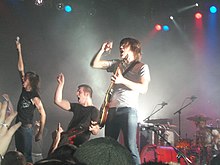 A heavy metal rock band on stage wearing black or white T-shirts and jeans, in the spotlight performing before an audience.