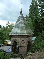 A square sandstone structure with an arch on each side, closed by a railing. On its top is a slate spire.