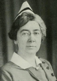 A middle-aged white woman with short greying hair, wearing a nurse's uniform and glasses