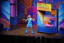 A distant stage shot of a female performer wearing bright clothes and singing into a headset microphone. A jack-in-the-box style puppet sits behind her on a movable section, which is brightly coloured.