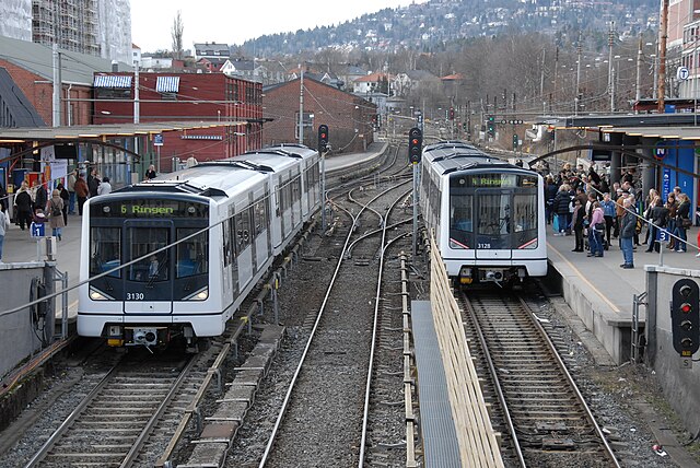 An open train station with three tracks and two side platforms filled with people. At each platform is parked a white three-car train.