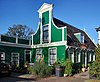 Bakstenen huis met ingezwenkt, door een fronton bekroond en door pilasters geleed houten voorschot. Links een vooruitspringende vleugel van hout met hoekvensters en een borstwering met pilasters