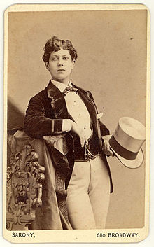 A white woman in menswear, holding a tophat