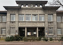 Cliché couleur. Bâtiment public. Rez-de-chaussée du corps central comprend un porche à deux colonnes encadré de deux fenêtres, au-dessus cinq fenêtres surmontées d'un fronton. Départ de deux ailes.