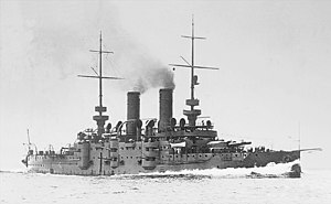 A large gray warship steams through the water, creating a large wave at the front.