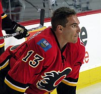 Hockey player in red uniform with his helmet off. He looks onto the ice.