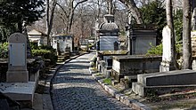 Farbfotografie eines Friedhofs mit mehreren alten Grabsteinen und Mausoleen, durch deren Mitte vertikal ein schmaler Pflasterweg führt. Im Hintergrund sind kahle Bäume.