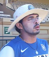 Young man wearing glasses and a white broad brimmed hat. He is wearing a black string necklace, blue T-shirt, and has a thin sideburn that links to a narrow beard, and a small thin moustache. He is wearing a light layer of white zinc cream on his face. A brick stand for spectators is in the background.