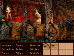 Ein Bild, das mit seiner niedrigen Farbtiefe wie ein Computerspiel-Screenshot aus den frühen 1990er-Jahren anmutet. Es zeigt einen Abenteurer bei der Bergung einer goldenen Götzenfigur aus einer Schatzhöhle. Im unteren Teil der Abbildung sind rechts verschiedene Gegenstandssymbole und links Verben wie „Nehmen“, „Benutzen“ und „Reden“ abgebildet. Der Mauszeiger deutet auf die Götzenfigur, um den aktuellen Befehl „Benutze Peitsche mit Götzenfigur“ zu erzeugen.