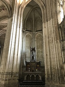 Haute chapelle ouverte sur deux côtés vers le déambulatoire. En bas de l'image, autel présentant des petites murailles sculptées et surmontée d'une femme vêtue de bleu et apprenant à une petite fille à lire.