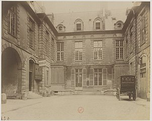 Cour de l'hôtel. On distingue au fond le signe d'une fabrique d'horlogerie installée dans l'ancien corps de logis, et, à gauche, celle d'une boutique de drapeaux. Photographie par Eugène Atget, 1901.