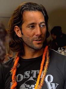 A bearded, long-haired man in a black shirt, with orange necklaces around his neck.