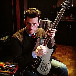 Josh Joplin wearing jeans and a sweater with collared shirt and tie underneath, sitting down, holding an electric guitar, and looking at camera over his glasses