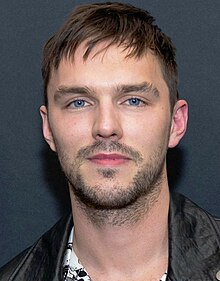 A young, Caucasian man with short, dark hair and facial stubble wearing a black shirt looks away from a microphone against a grey and blue background.