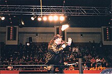 un homme habillé en noir et portant une casquette s'adresse au public depuis un ring.