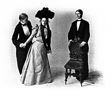 Three female actors, with two dressed as men and the third, in the center, dressed in an elegant gown with a large headpiece; the leftmost character offers a chair, being held by the rightmost character, to the woman in the center.